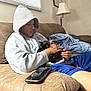 person, hoodie, kitten, black_cat, couch, blanket, smartphone, hand, window, blind, indoor, casual_clothing, relaxing, seated, brown_furniture, portrait, young_adult, comfort, pet, home