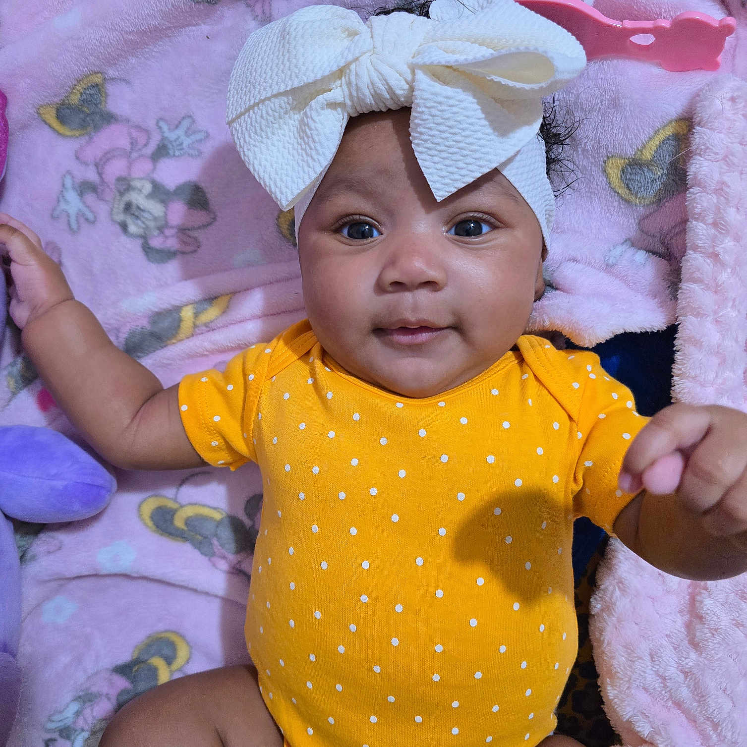 Keivelyn is registered to the contest to win money with this photo: baby, bow, cartoon_characters, child, closeup, cute, face, hand, happy, headband, indoors, infant, laying_down, pink_blanket, polka_dot, portrait, smiling, soft_texture, toddler, yellow_onesie