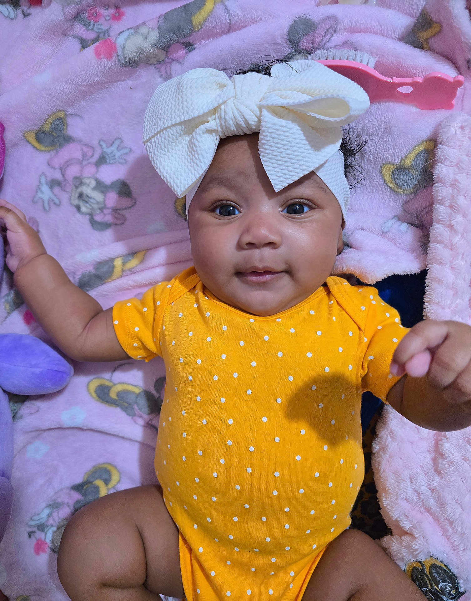 Keivelyn is registered to the contest to win money with this photo: baby, bow, cartoon_characters, child, closeup, cute, face, hand, happy, headband, indoors, infant, laying_down, pink_blanket, polka_dot, portrait, smiling, soft_texture, toddler, yellow_onesie