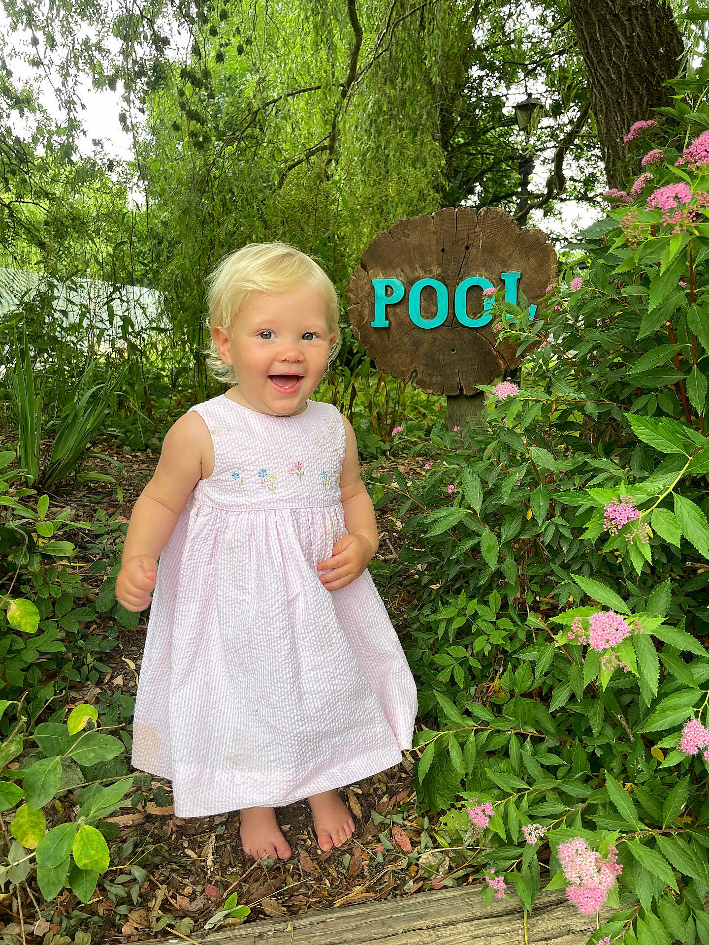Indigo joined the competition — help win amazing prizes! child, toddler, dress, barefoot, garden, flowers, greenery, plants, tree, wooden_sign, outdoor, happy, smiling, nature, summer, daylight, leaves, path, wood, pink