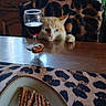 cat, wine_glass, pretzels, breadsticks, plate, table, chair, kitchen, leopard_print, tablecloth, curious, wooden_furniture, window, curtains, sink, bottle, food, snack, indoor, pet