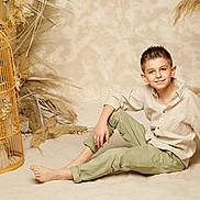 Timoté a rejoint le concours — aidez-le/la à gagner de superbes lots ! child, boy, smiling, barefoot, sitting, sand, birdcage, dried_plants, beige, earth_tones, portrait, casual_clothing, indoor, decor, natural_light, relaxed, young, short_hair, buttoned_shirt, green_pants