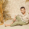 barefoot, beige, birdcage, boy, buttoned_shirt, casual_clothing, child, decor, dried_plants, earth_tones, green_pants, indoor, natural_light, portrait, relaxed, sand, short_hair, sitting, smiling, young