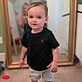 toddler, child, barefoot, black_tshirt, shorts, mirror, reflection, indoor, wooden_floor, person, home, blurred_background, photography, standing, casual_clothing, young_child, portrait, smiling, short_hair, cute