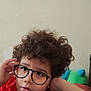 child, curly_hair, red_shirt, glasses, freckles, face, casual, indoors, couch, colorful_cushions, relaxed, portrait, young, hand_on_head, expression, brown_hair, light_skin, wall, home, person