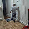 Noah is registered to the contest to win money with this photo: back_view, bowl, broom, child, curious, dim_light, door, dustpan, exploring, floor, footwear, hallway, hat, home, indoor, pajamas, person, plaid, wall, wood_floor