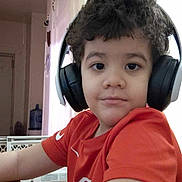 Noah joined the competition — help win amazing prizes! child, headphones, curly_hair, red_shirt, indoor, table, water_jug, baby_gate, door, wall, person, sitting, relaxed, home, furniture, casual, ear_covers, portrait, young, technology