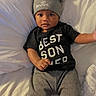 Asher is registered to the contest to win money with this photo: baby, child, infant, hat, gray_hat, bear_ears, black_shirt, text_on_shirt, lying_down, bed, white_bed, pants, gray_pants, cute, portrait, indoor, person, young_child, looking_at_camera, comfortable