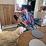 Mandie joined the competition — help win amazing prizes! man, wheelchair, dog, white_dog, indoor, kitchen, table, placemats, wood_paneling, window, decorations, chair, floor, smiling, jacket, casual_clothing, interaction, pet, happy, home