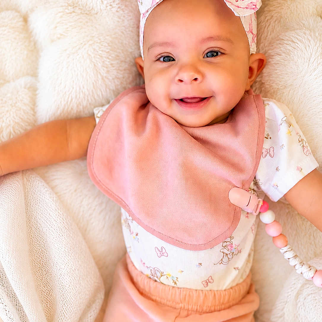 Amira is registered to the contest to win money with this photo: baby, smiling, headband, bib, pink_clothing, pacifier_clip, blanket, cute, infant, happy, face, child, soft_texture, pastel_colors, lying_down, portrait, adorable, clothing, beads, young_child