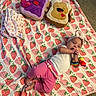 baby, blanket, strawberry_pattern, headband, plush_toy, peanut_butter_toast, jelly_toast, pink_pants, white_shirt, wrist_rattle, carpet, infant, cute, lying_down, indoor, child, playful, soft_toys, colorful, cozy