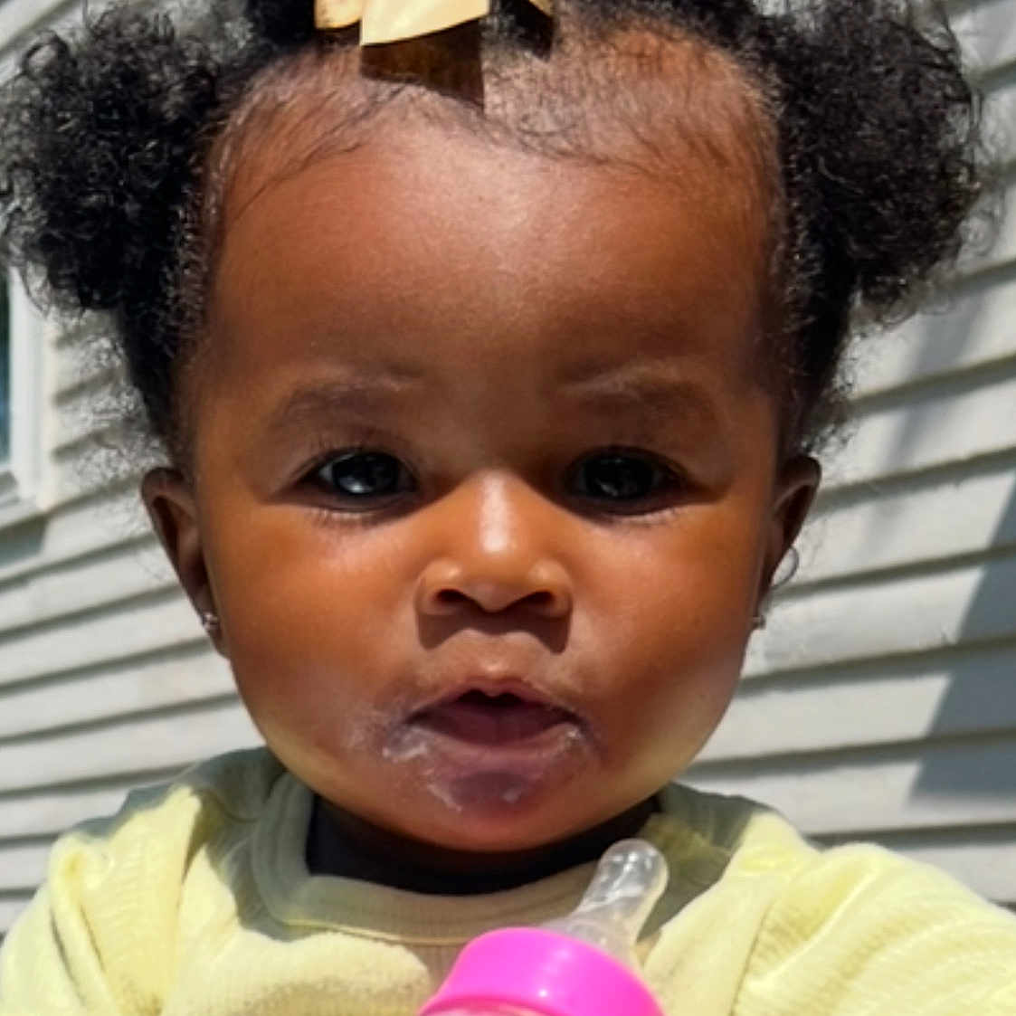 Amara joined the competition — help win amazing prizes! baby, child, clothing, curly_hair, cute, expression, face, feeding, holding, infant, milk, milk_bottle, outdoor, person, pink, portrait, siding, sunlight, yellow_bow, young_child