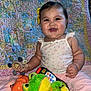 baby, child, smiling, happy, toy, stuffed_animal, teething_toy, quilt, yellow_bow, floral_top, pink_pants, indoor, candid, portrait, cute, infant, playful, colorful, soft_toys, remote_control