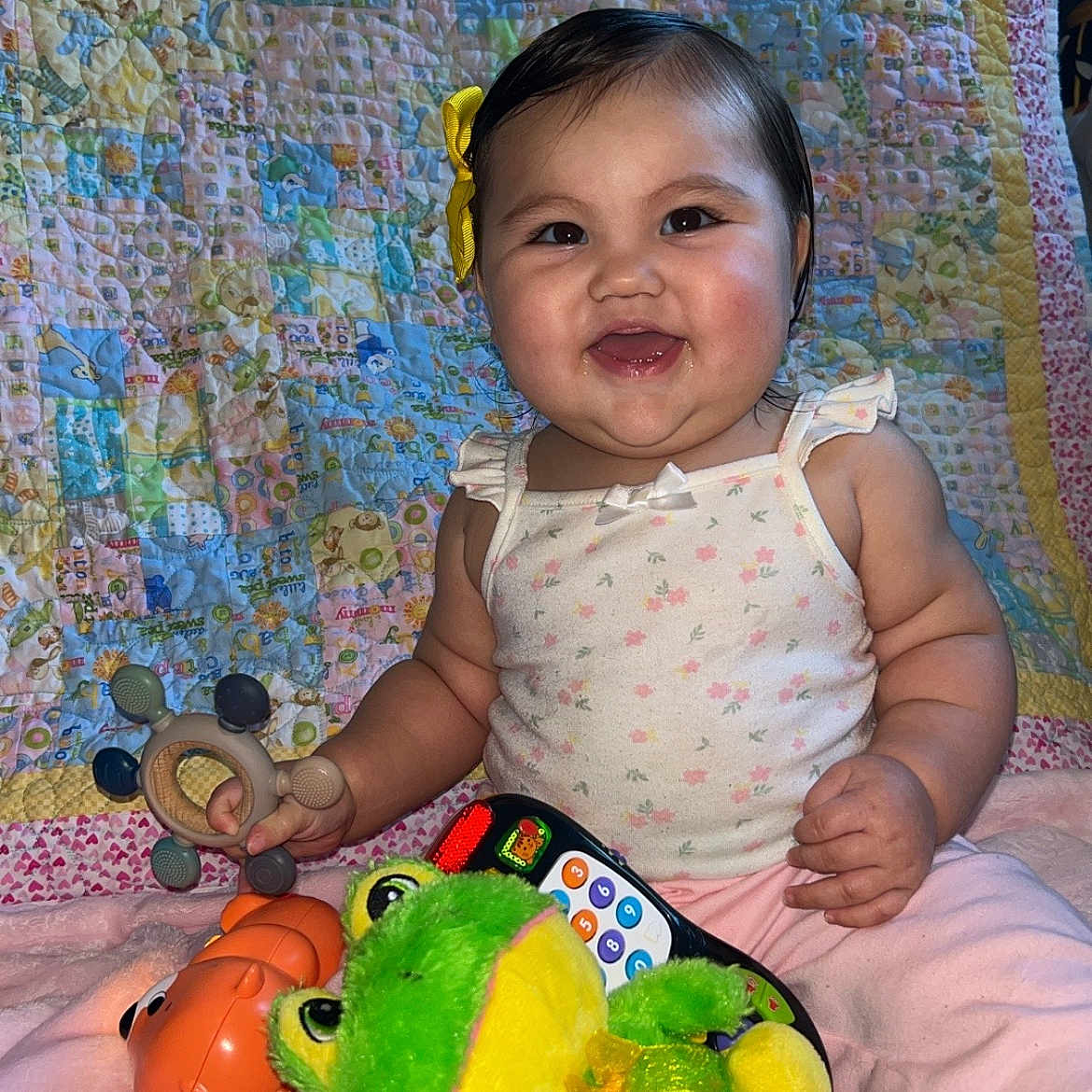 Annaleia joined the competition — help win amazing prizes! baby, candid, child, colorful, cute, floral_top, happy, indoor, infant, pink_pants, playful, portrait, quilt, remote_control, smiling, soft_toys, stuffed_animal, teething_toy, toy, yellow_bow