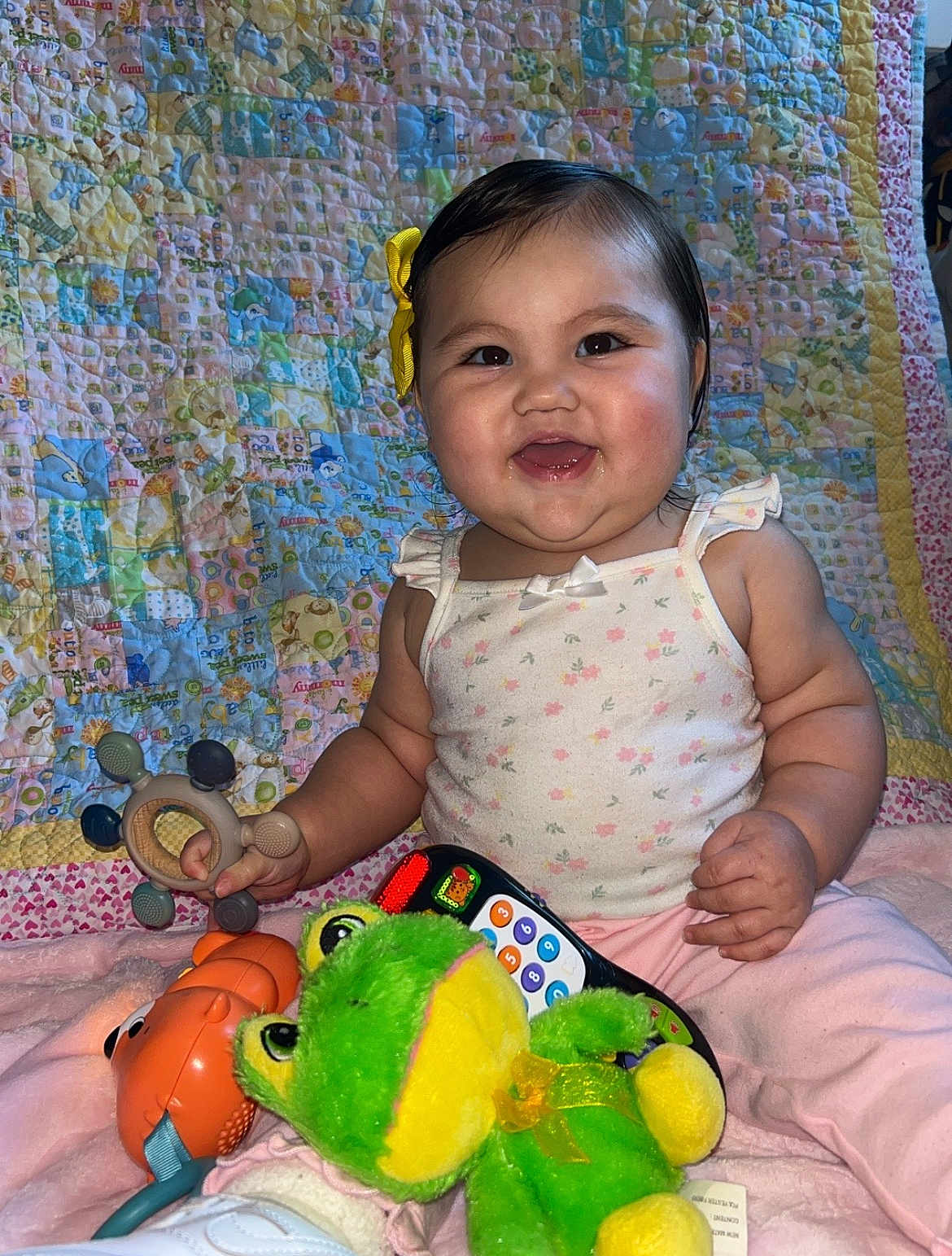 Annaleia joined the competition — help win amazing prizes! baby, child, smiling, happy, toy, stuffed_animal, teething_toy, quilt, yellow_bow, floral_top, pink_pants, indoor, candid, portrait, cute, infant, playful, colorful, soft_toys, remote_control