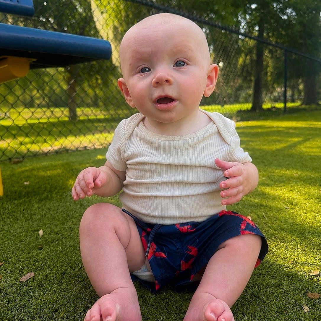 Christopher joined the competition — help win amazing prizes! baby, child, grass, outdoor, sunlight, park, shirt, shorts, barefoot, face, expression, nature, fence, summer, sitting, person, cute, curious, skin, daytime