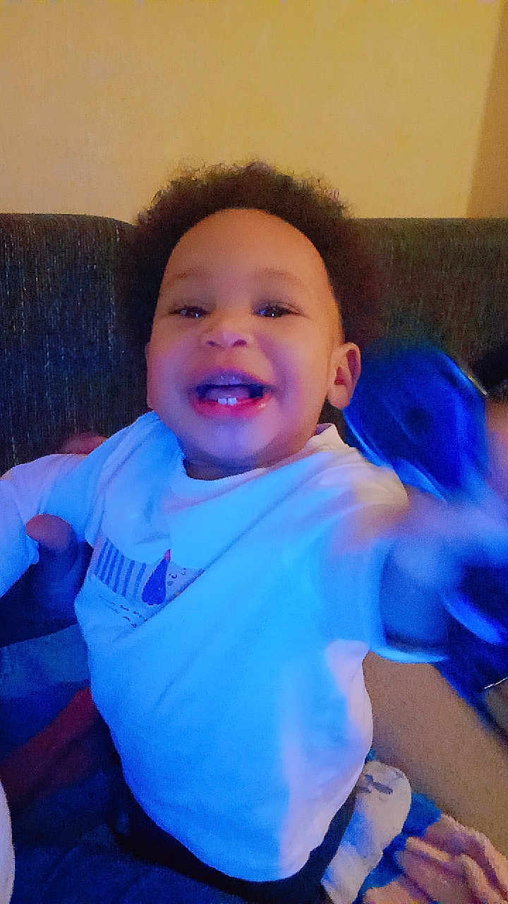 Nissam a rejoint le concours — aidez-le/la à gagner de superbes lots ! child, toddler, smile, teeth, afro, curly_hair, shirt, sitting, couch, indoors, portrait, happy, hand, arm, blurry_motion, toy, blue_tone, evening, baby, person