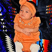 Lexi joined the competition — help win amazing prizes! baby, infant, child, portrait, sitting, orange_outfit, turban, headband, dress, embroidered_clothing, white_socks, blanket, patterned_background, textile, indoor, front_facing, serious_expression, hands_folded, colorful, photography