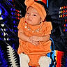 baby, infant, child, portrait, sitting, orange_outfit, turban, headband, dress, embroidered_clothing, white_socks, blanket, patterned_background, textile, indoor, front_facing, serious_expression, hands_folded, colorful, photography