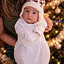 baby, child, christmas_tree, cozy, cute, festive, hat, holding, holiday_lights, indoor, infant, long_hair, ornaments, person, pom_pom, portrait, tinsel, warm_lighting, white_clothing, winter_wear