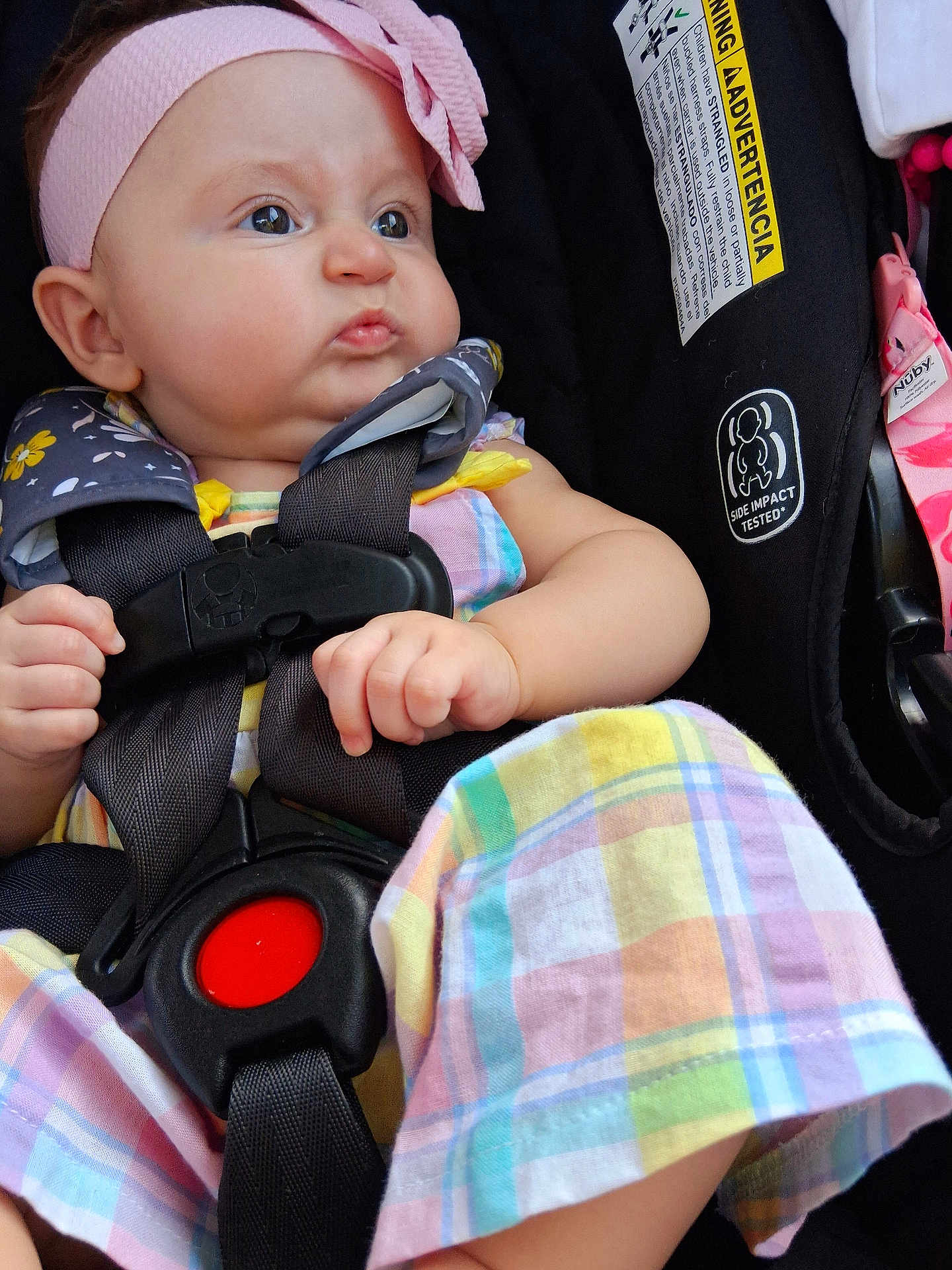 Brooklyn is registered to the contest to win money with this photo: baby, child, car_seat, headband, bow, plaid_clothing, pout, infant, seatbelt, safety_label, colorful, cute, expression, strapped_in, indoors, close_up, person, young_child, hand, face