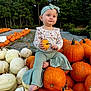 autumn, baby, barefoot, child, curious_expression, flared_pants, green_headband, headband, holding, long_sleeve_shirt, nature, orange_pumpkin, outdoor, pants, patterned_shirt, pumpkin, sitting, toddler, trees, white_pumpkin