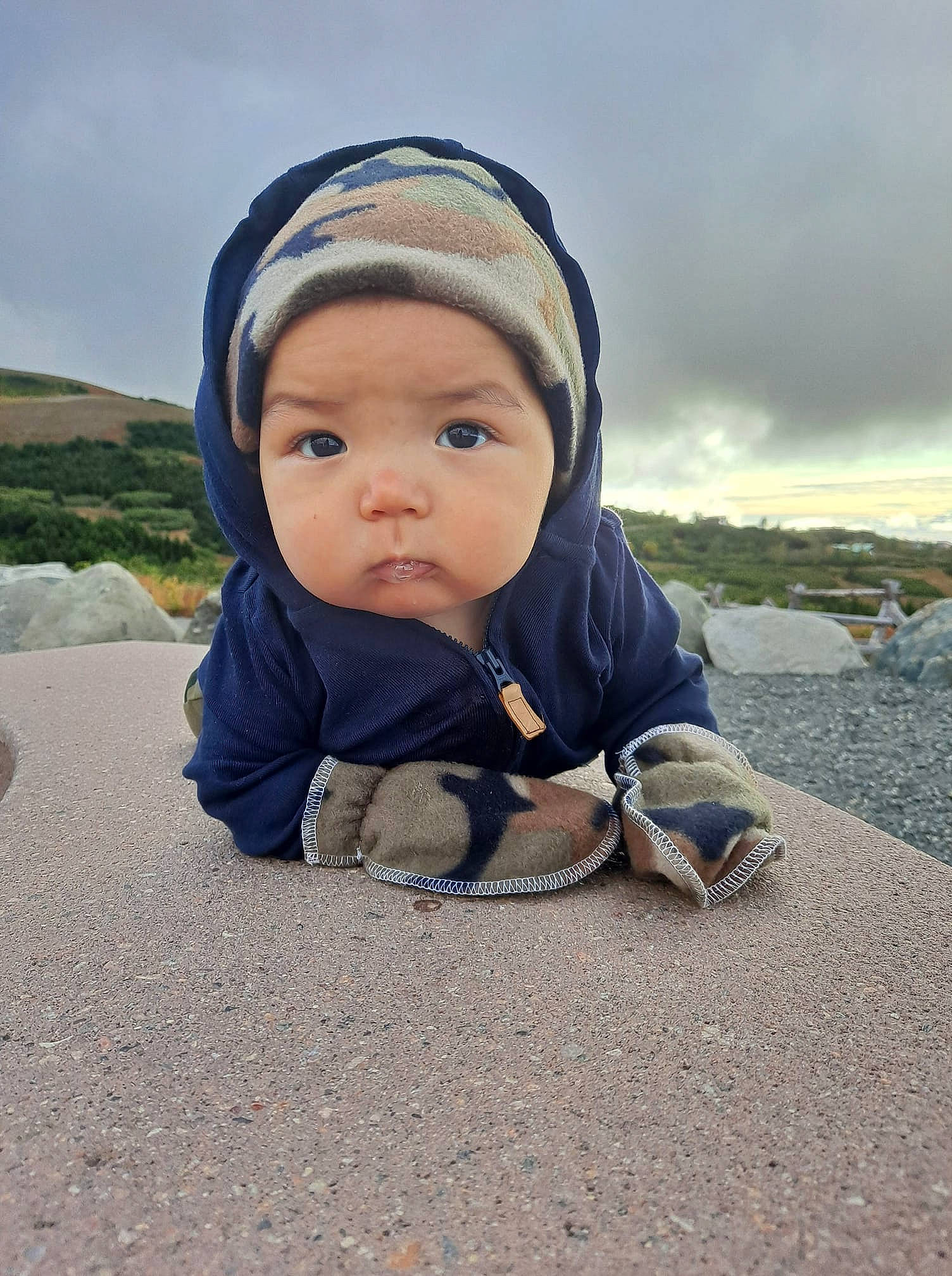Junior is registered to the contest to win money with this photo: asphalt, baby, cap, cloud, comfort, freezing, grass, happy, headwear, jacket, landscape, leisure, people_in_nature, person, road_surface, rock, sand, sitting, sky, toddler