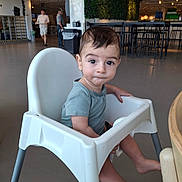 John is registered to the contest to win money with this photo: toddler, child, high_chair, indoor, cafeteria, person, furniture, table, floor, shirt, footwear, expression, curious, hair, skin, legs, seat, modern, plant_wall, people