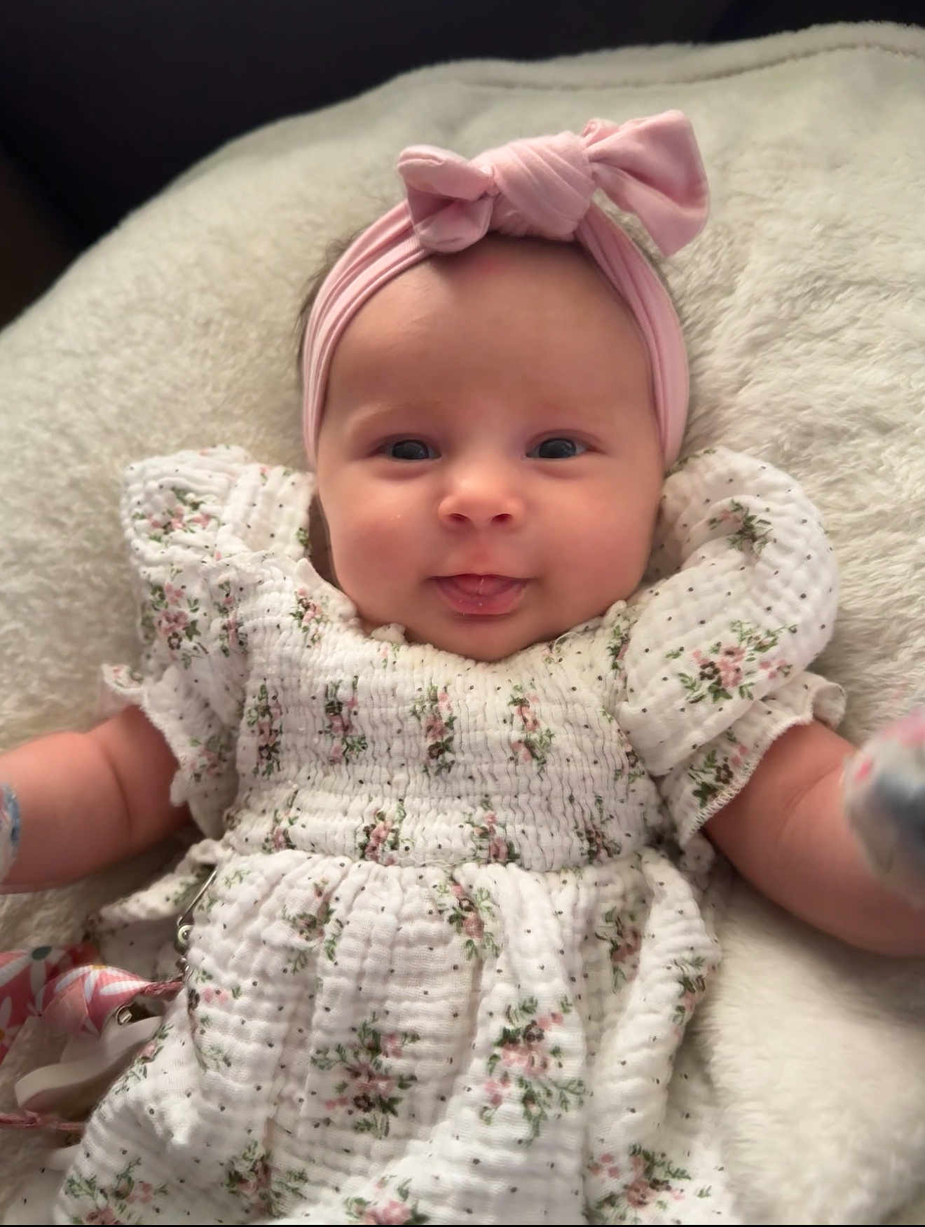 Scarlett is registered to the contest to win money with this photo: baby, infant, girl, headband, pink, tongue_out, floral_dress, white_dress, smiling, cute, soft_blanket, lying_down, portrait, child, adorable, closeup, happy, indoor, person, baby_clothes