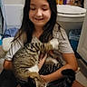 child, cat, bathroom, toilet, towel, shampoo_bottle, floor, smiling, long_hair, gray_tshirt, sitting, pet, playful, indoor, young_girl, tabby_cat, holding, legs_crossed, toiletries, trash_can
