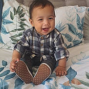 Kai joined the competition — help win amazing prizes! baby, child, smiling, plaid_shirt, sitting, bed, pillow, headboard, footwear, shoe, pattern, leaf_print, indoor, happy, person, infant, cute, cozy, home, portrait
