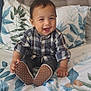 baby, child, smiling, plaid_shirt, sitting, bed, pillow, headboard, footwear, shoe, pattern, leaf_print, indoor, happy, person, infant, cute, cozy, home, portrait