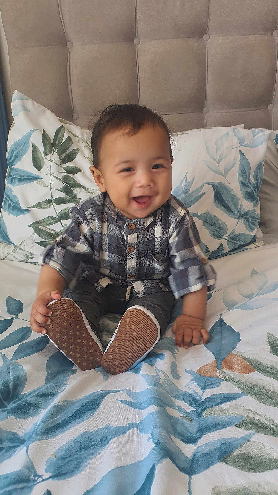 Kai joined the competition — help win amazing prizes! baby, child, smiling, plaid_shirt, sitting, bed, pillow, headboard, footwear, shoe, pattern, leaf_print, indoor, happy, person, infant, cute, cozy, home, portrait