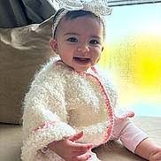 Lilia participe au concours pour gagner de l'argent avec cette photo : baby, child, smiling, headband, bow, white_jacket, pink_trim, couch, window, natural_light, indoors, cute, happy, portrait, infant, sitting, cozy, warm_clothing, person, face