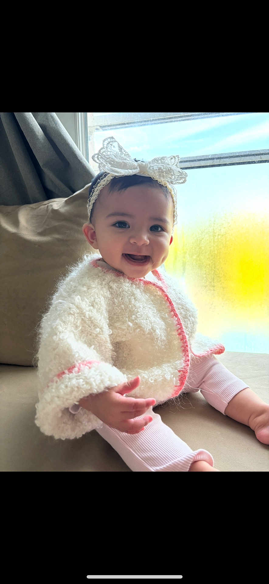 Lilia participe au concours pour gagner de l'argent avec cette photo : baby, child, smiling, headband, bow, white_jacket, pink_trim, couch, window, natural_light, indoors, cute, happy, portrait, infant, sitting, cozy, warm_clothing, person, face