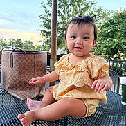 Melanie joined the competition — help win amazing prizes! baby, child, smile, yellow_outfit, table, metal_table, bag, coffee_cup, umbrella, outdoor, greenery, trees, sunset, happy, sitting, cute, portrait, person, daylight, casual