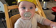 Remi joined the competition — help win amazing prizes! baby, high_chair, pacifier, hat, bee_hat, child, indoor, restaurant, wooden_chair, table, person, face, clothing, curious, seated, casual, people, background, lighting, cute
