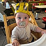 baby, high_chair, pacifier, hat, bee_hat, child, indoor, restaurant, wooden_chair, table, person, face, clothing, curious, seated, casual, people, background, lighting, cute