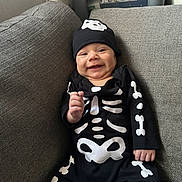 Remi is registered to the contest to win money with this photo: baby, smiling, costume, skeleton, hat, black_clothing, couch, gray, infant, indoors, happy, cute, child, portrait, seated, clothing, small_hand, face, young, cozy