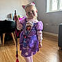 Aria is registered to the contest to win money with this photo: child, girl, costume, purple_dress, cat_ears, face_paint, broom, indoor, wooden_floor, window, natural_light, playful, smiling, person, toy, dress_up, headband, flooring, furniture, cute