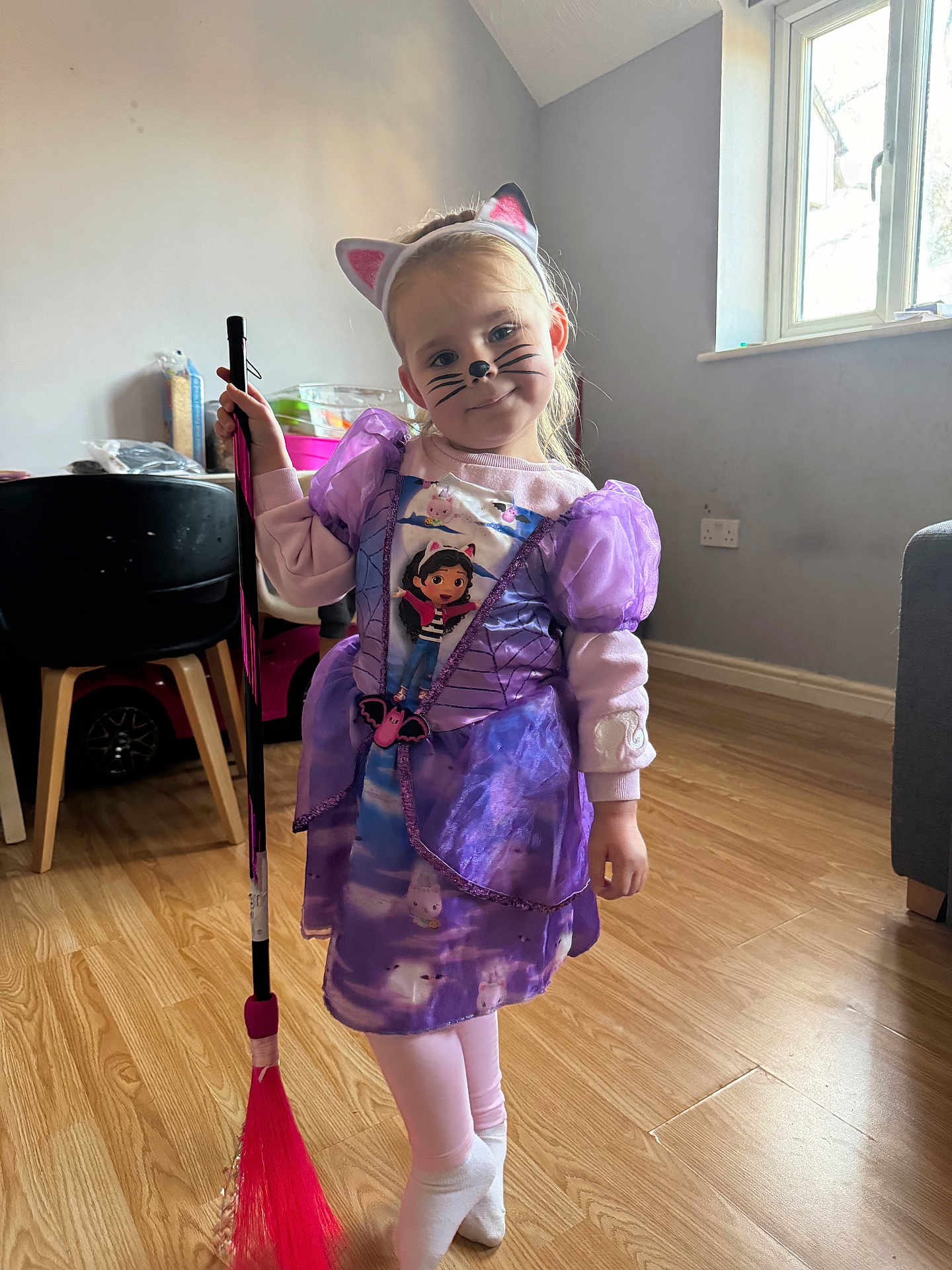 Aria is registered to the contest to win money with this photo: child, girl, costume, purple_dress, cat_ears, face_paint, broom, indoor, wooden_floor, window, natural_light, playful, smiling, person, toy, dress_up, headband, flooring, furniture, cute