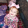 Rosalie is registered to the contest to win money with this photo: toddler, child, pink_bow, plaid_blanket, stuffed_animal, purple_blanket, pink_dress, indoor, smiling, happy, face, person, soft_toys, cozy, baby, cute, seated, clothing, blanket, home