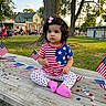 american_flag, casual, celebration, child, confetti, curly_hair, cute, daylight, event, grass, outdoor, park, patriotic_clothing, people, picnic_table, pink_bow, sitting, stars_and_stripes, toddler, tree