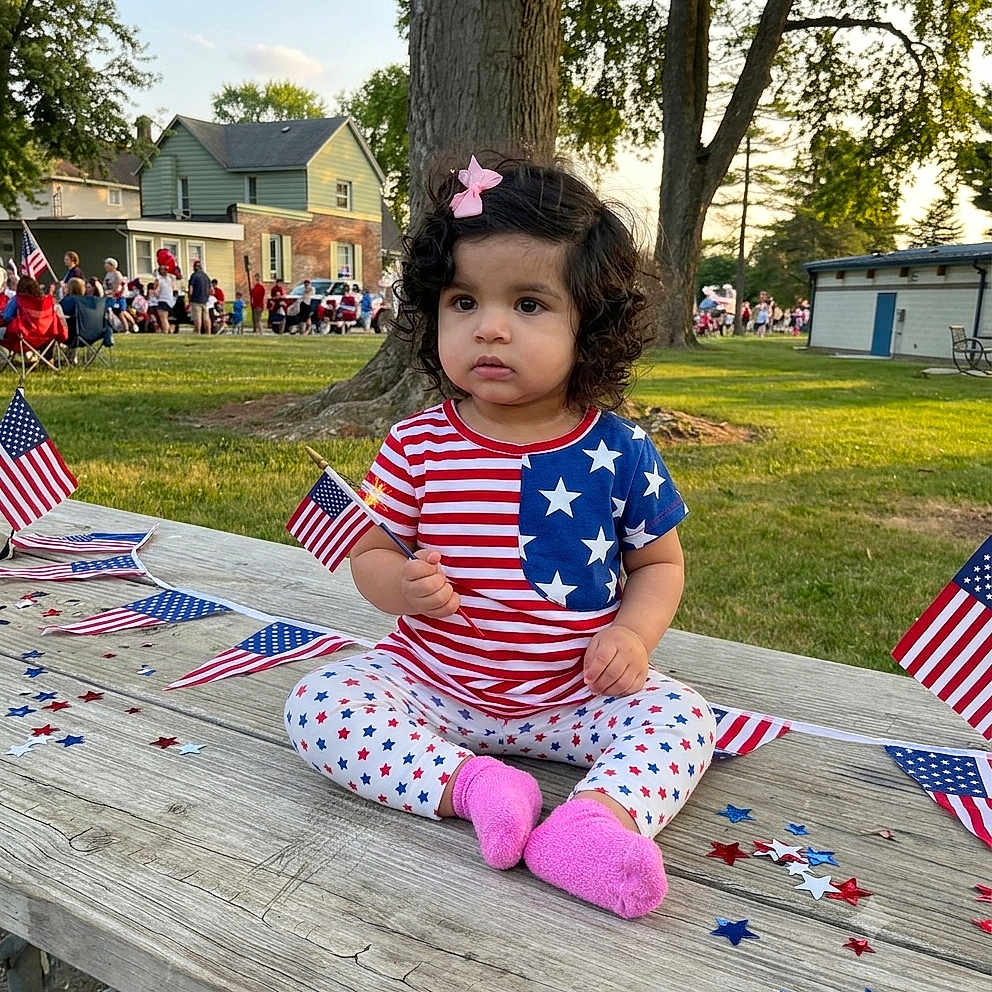 Kshaira joined the competition — help win amazing prizes! toddler, child, curly_hair, pink_bow, american_flag, patriotic_clothing, stars_and_stripes, picnic_table, confetti, outdoor, park, grass, tree, people, daylight, casual, sitting, celebration, event, cute