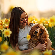 Vanille a rejoint le concours — aidez-le/la à gagner de superbes lots ! woman, dog, flower_field, daffodils, yellow_flowers, outdoor, spring, sunlight, nature, pet, portrait, happy, smiling, long_hair, casual_clothing, greenery, warm_light, affection, companionship, close_up