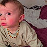 baby, beaded_necklace, bed, beige_sweater, child, cozy, crawling, curious_expression, face, hair, hand, home, indoor, person, photo_wall, pillow, plush_blanket, purple_blanket, rosy_cheeks, young_child