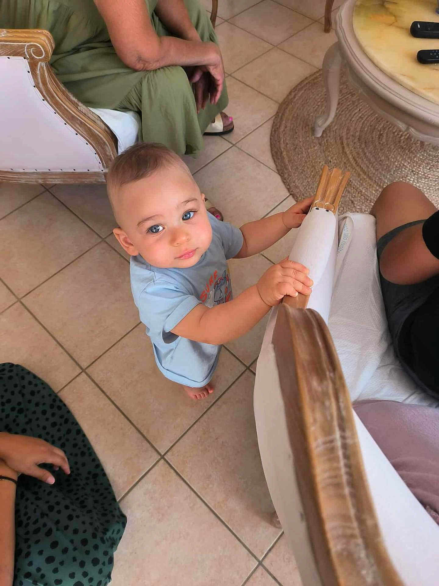 Aron participe au concours pour gagner de l'argent avec cette photo : toddler, child, indoor, curious, blue_eyes, wooden_chair, tile_floor, people, hand, foot, sitting, clothing, adult, table, remote_control, carpet, casual, furniture, family, home
