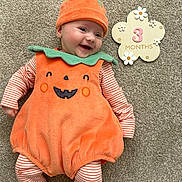 Clementine is registered to the contest to win money with this photo: baby, infant, pumpkin_costume, orange, green, striped_clothing, smiling, lying_down, carpet, 3_months_sign, flower_shape, cute, happy, holiday_costume, child, portrait, festive, indoors, costume, seasonal