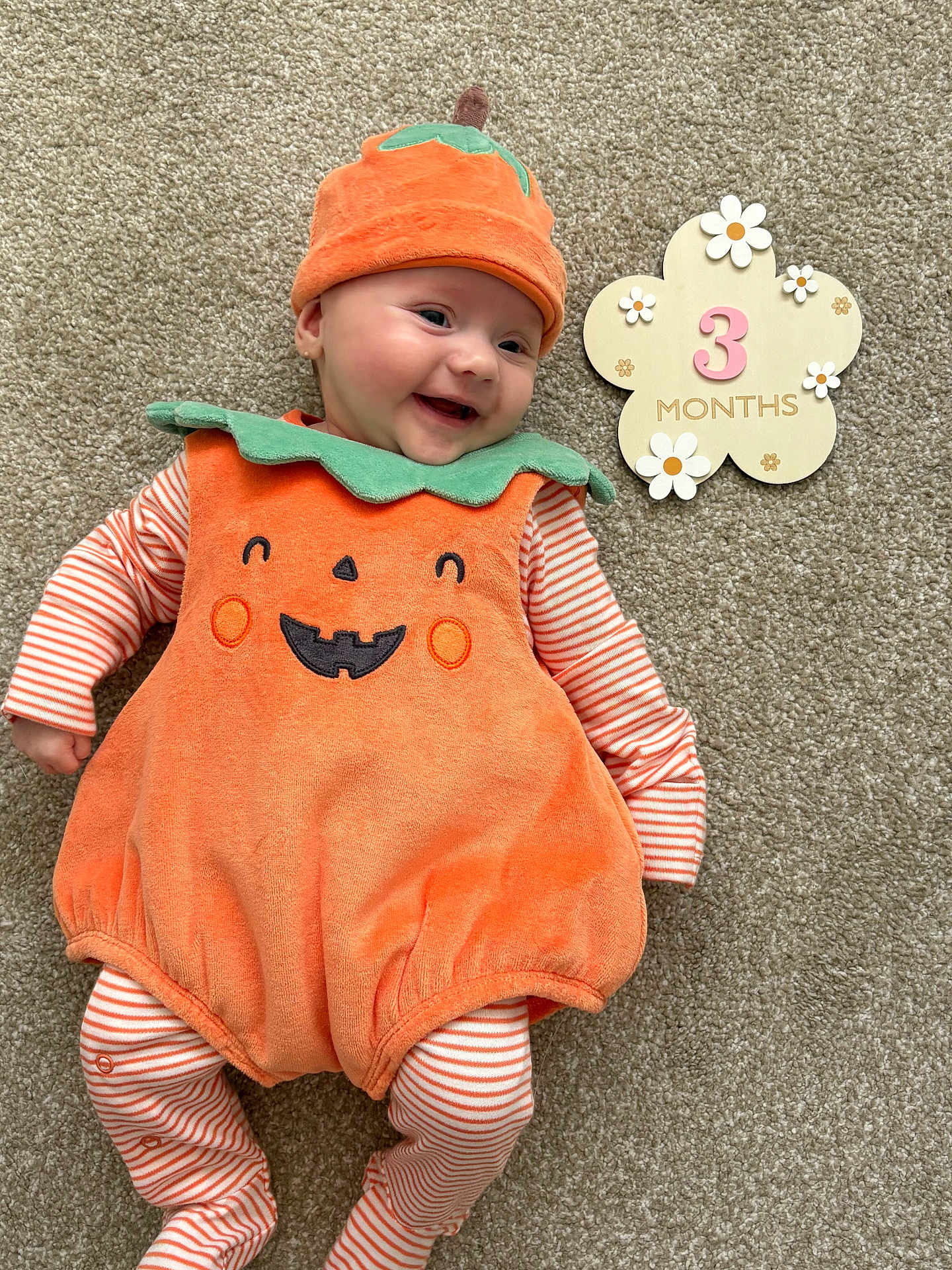 Clementine is registered to the contest to win money with this photo: baby, infant, pumpkin_costume, orange, green, striped_clothing, smiling, lying_down, carpet, 3_months_sign, flower_shape, cute, happy, holiday_costume, child, portrait, festive, indoors, costume, seasonal