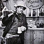 bar, beard, black_and_white, bottles, costume, cowboy, dog, facial_hair, hat, hat_on_dog, indoors, man, mug, old_west, portrait, rifle, saloon, small_dog, vintage, wooden_wall